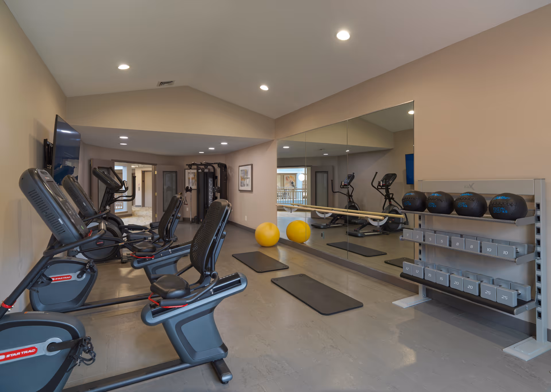 Bright indoor fitness room with recumbent bikes, exercise mats, medicine balls and weights against a mirrored wall.