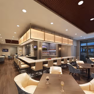 Modern dining room and bar area with upholstered chairs, tables, wood flooring, and warm overhead lighting.