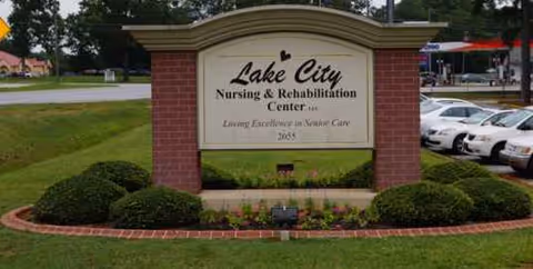Lake City Nursing and Rehabilitation Center sign on brick pillars with landscaping and parked cars.