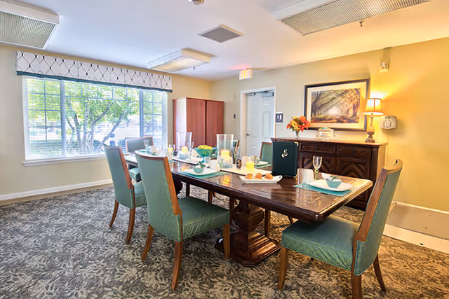 A bright dining room with a large wooden table set for six people with plates, glasses, and candles. The room has green upholstered chairs, a large window with a view of trees outside, a sideboard with a lamp and framed artwork on the wall, and a carpeted floor.