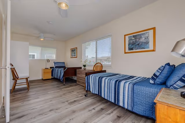 A bright and clean bedroom with two single beds, each with blue striped bedding and pillows. The room has light-colored walls, two windows with blinds, wooden furniture including a nightstand, a chair, and a small desk with a chair. There are framed pictures on the walls and a ceiling fan above.