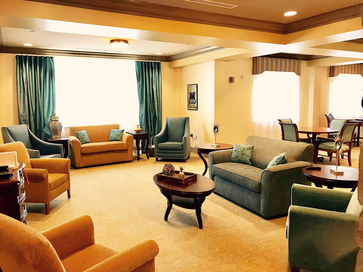 A spacious and well-lit living room area with multiple upholstered chairs and sofas in shades of blue and mustard yellow. There are wooden coffee and side tables with decorative items placed on them. Large windows with teal curtains and valances allow natural light to fill the room. In the background, there is a dining area with tables and chairs near windows with sheer curtains.