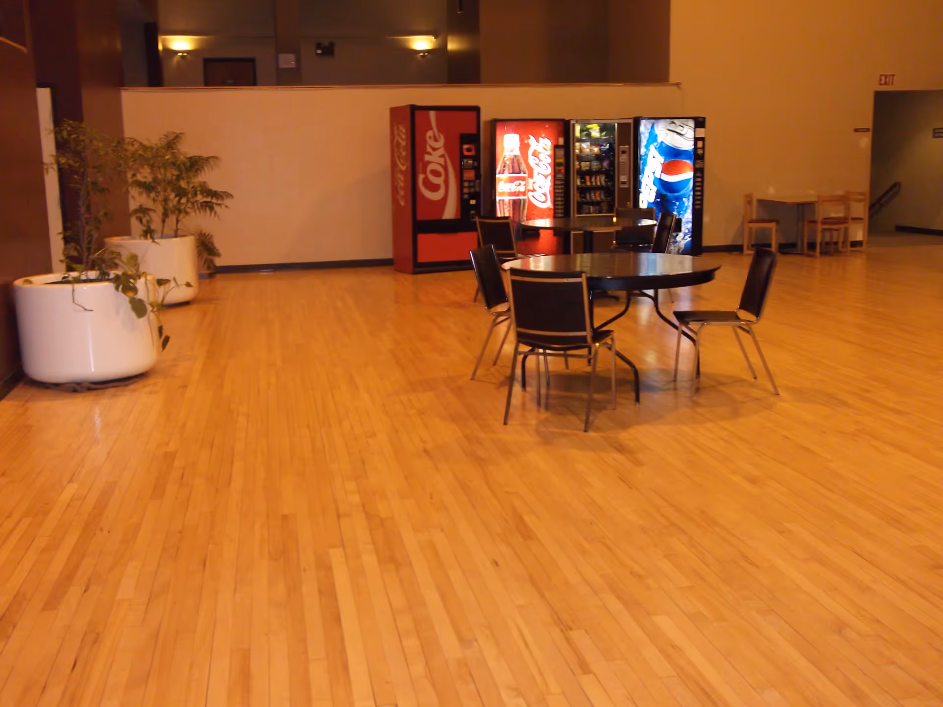A spacious indoor area with a polished wooden floor featuring a round table with four chairs in the center. In the background, there are three vending machines for Coke, snacks, and Pepsi. Large white planters with green plants are placed along the left wall, and there is an exit door visible on the right side.