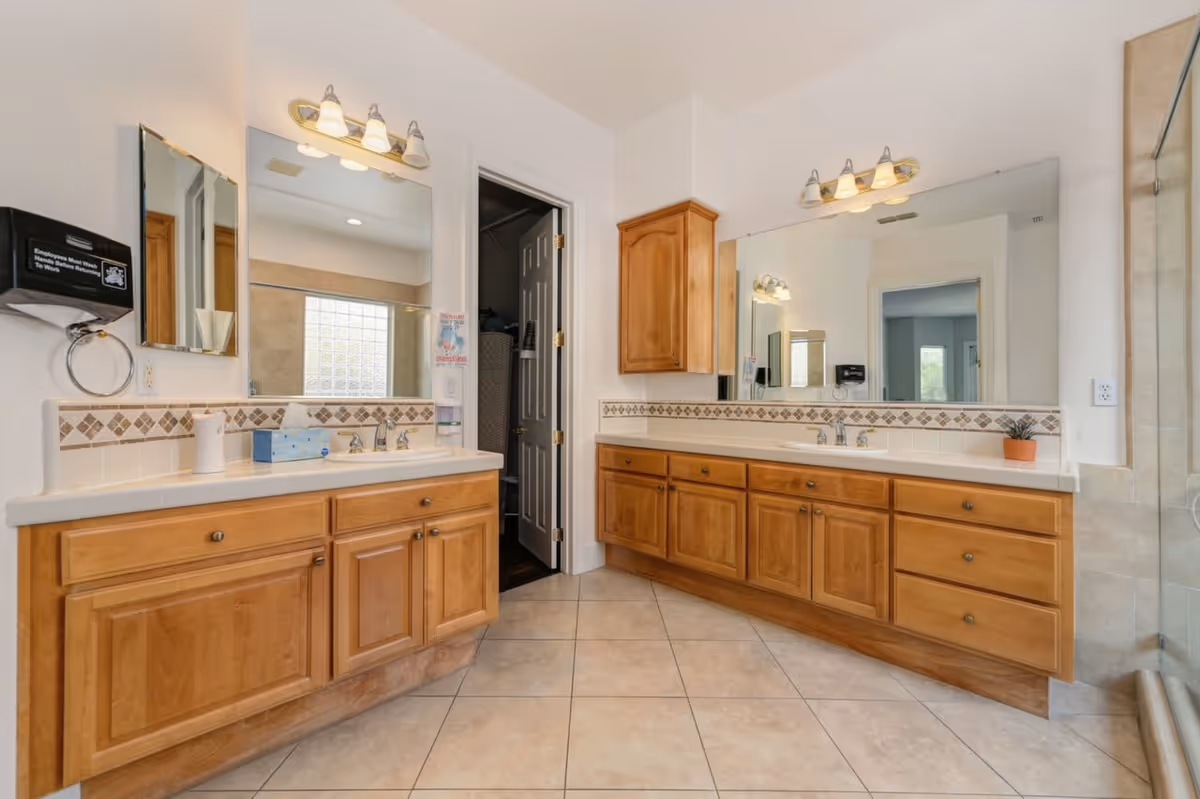 A spacious bathroom with two wooden vanities featuring multiple drawers and cabinets, each with a large mirror and overhead lighting. The floor is tiled, and there is a glass-enclosed shower visible on the right side. A door in the center leads to another room, and there is a soap dispenser and paper towel holder mounted on the wall.