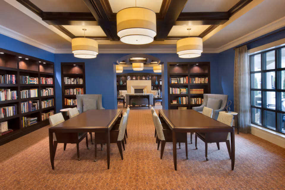 A well-lit library or reading room with blue walls, dark wooden bookshelves filled with books, two long wooden tables with chairs, and two upholstered armchairs near a large window with curtains. The ceiling has dark wooden beams and three large round pendant lights. In the background, there is an adjoining room with more seating and a fireplace.
