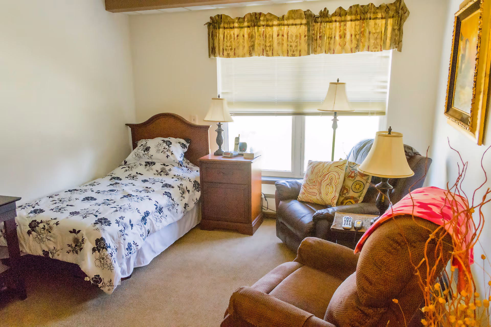 Small bedroom with a single bed, bedside table, two armchairs and lamps beside a large window.