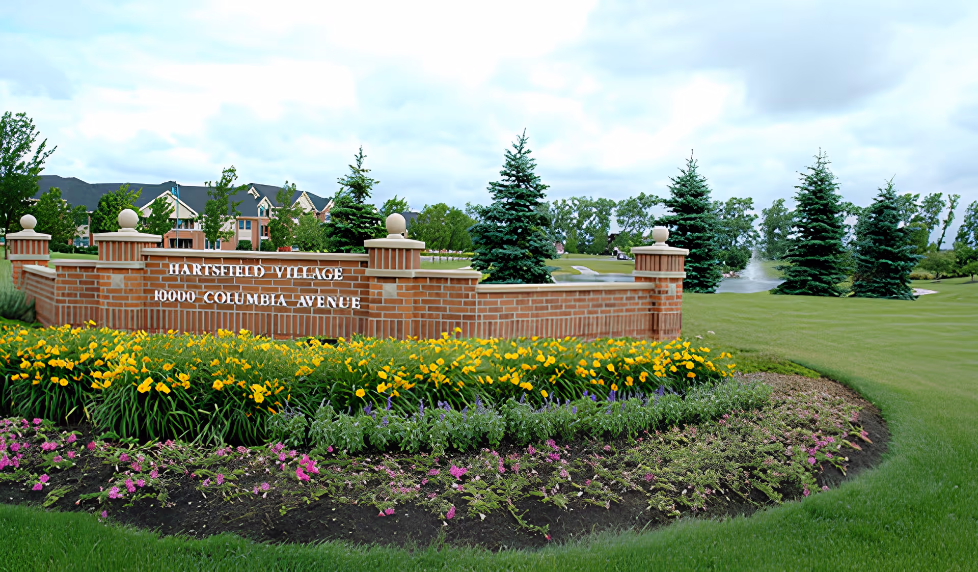 Brick entrance sign for Hartsfield Village with the address 10000 Columbia Avenue, surrounded by well-maintained flower beds with yellow and purple flowers, green grass, and several evergreen trees in the background under a cloudy sky.