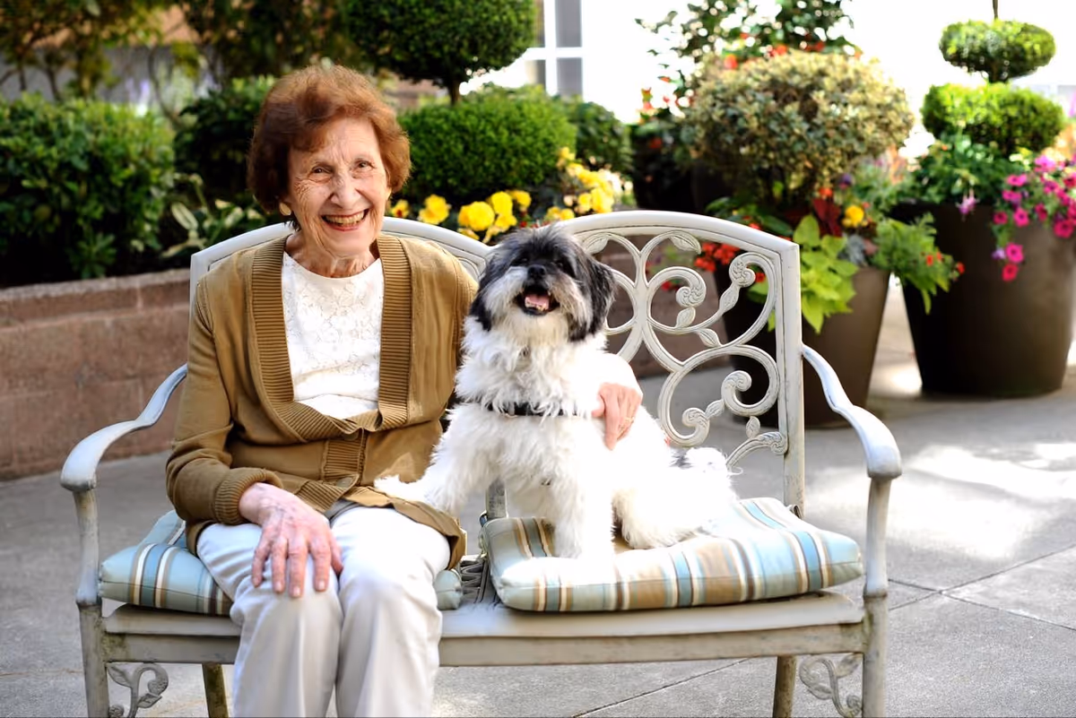 An elderly woman with short curly hair, wearing a brown cardigan and white pants, sitting on a cushioned metal bench outdoors. She is smiling and has her arm around a small black and white dog sitting next to her. Behind them are large potted plants with green foliage and colorful flowers.