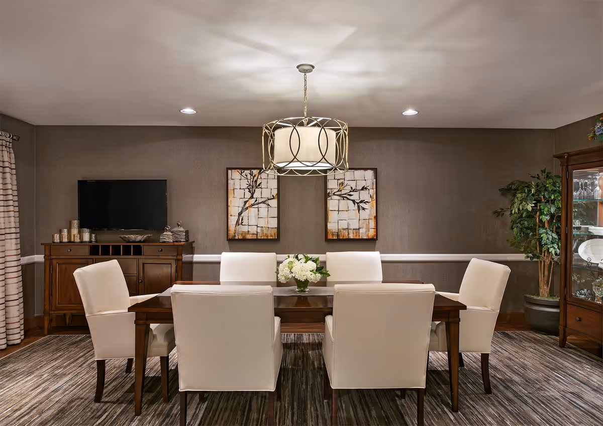 A dining room with a rectangular wooden table surrounded by six white upholstered chairs. A decorative chandelier hangs above the table, which has a floral centerpiece. The room features a patterned carpet, a wooden cabinet with a TV on top, two framed artworks on the wall, a glass display cabinet with dishes, and a large potted plant in the corner.