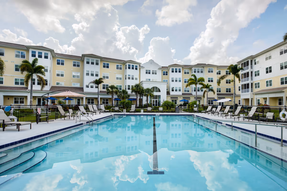 Outdoor swimming pool with clear blue water reflecting the sky and clouds, surrounded by lounge chairs, umbrellas, palm trees, and a multi-story residential building in the background under a partly cloudy sky.