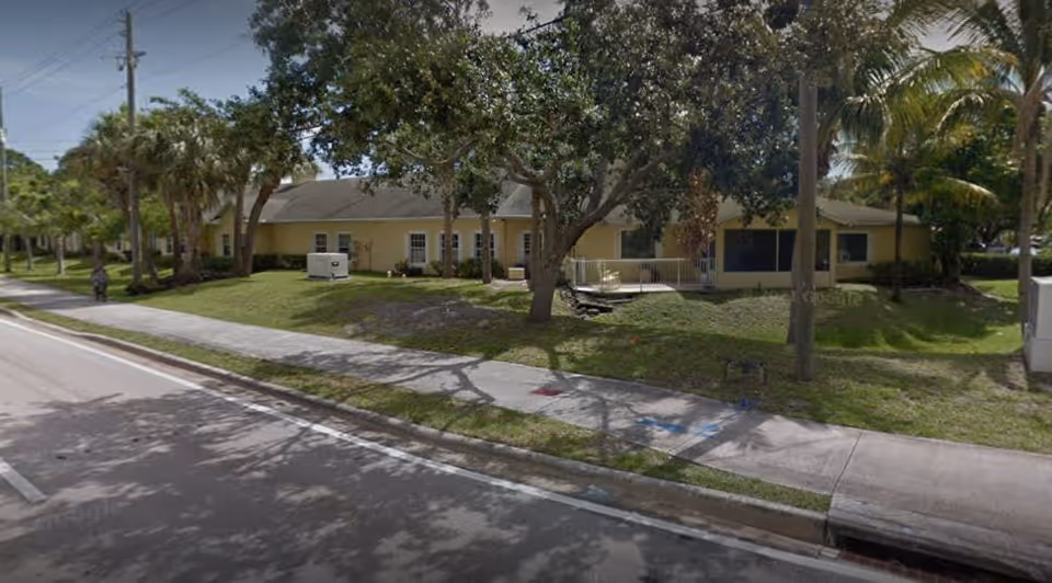 Single-story yellow building with lawn and trees fronting a sidewalk and street.