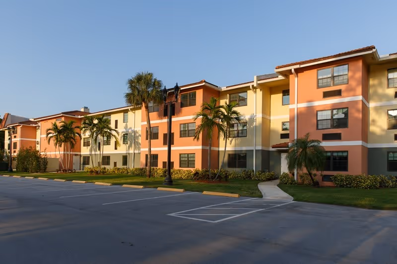 Exterior view of a three-story senior living facility building with a colorful facade in shades of orange, yellow, and beige. The building is surrounded by palm trees and landscaped greenery, with a parking lot in the foreground under a clear blue sky.