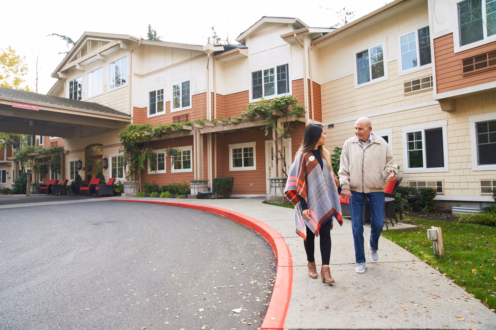 An elderly man and a younger woman walking and talking on a sidewalk outside a senior living facility building with beige and brown exterior walls, windows, and greenery around.