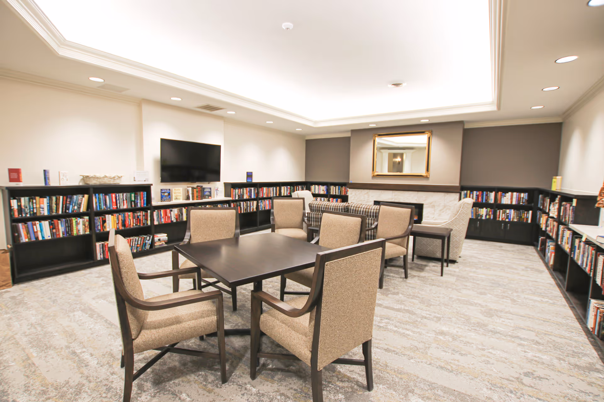 A cozy library room with beige carpet and neutral walls. There are several bookshelves filled with books lining the walls. In the center, there is a dark wooden table surrounded by six beige upholstered chairs. At the back of the room, there is a seating area with a plaid sofa, an armchair, a side table, and a fireplace with a gold-framed mirror above it. A flat-screen TV is mounted on the wall above one of the bookshelves.