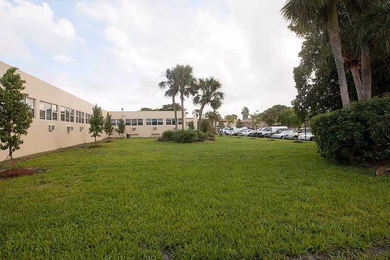 A beige low-rise building with a green lawn, palm trees, and a parking lot.