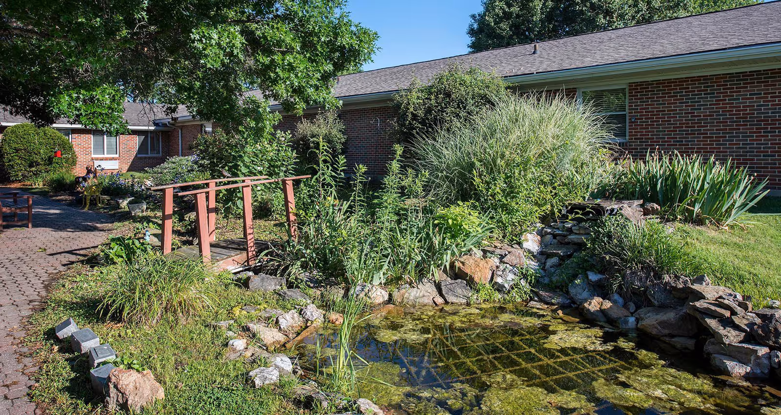 A landscaped outdoor area at Rolla Presbyterian Manor featuring a small wooden bridge over a pond surrounded by rocks and lush greenery, with a brick building and windows in the background under a clear blue sky.