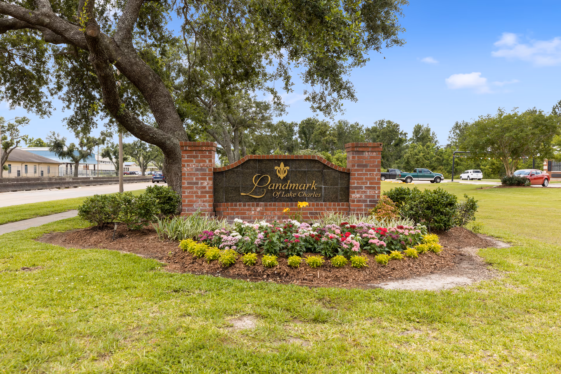 A landscaped entrance sign for Landmark of Lake Charles, featuring a brick and stone structure with the facility name in gold lettering, surrounded by colorful flowers and greenery under a blue sky.