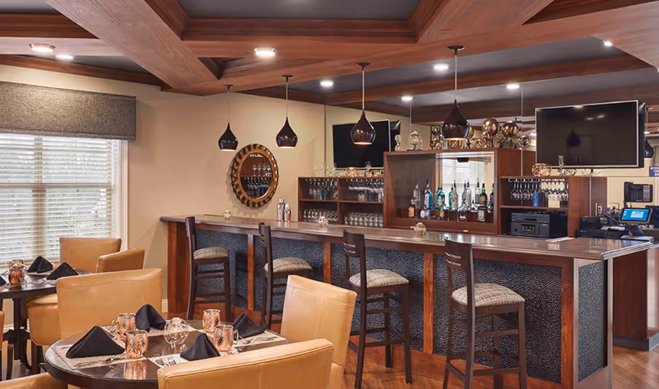 Interior dining and bar area with a wooden coffered ceiling, a long bar with stools, and tables set with napkins and glassware.