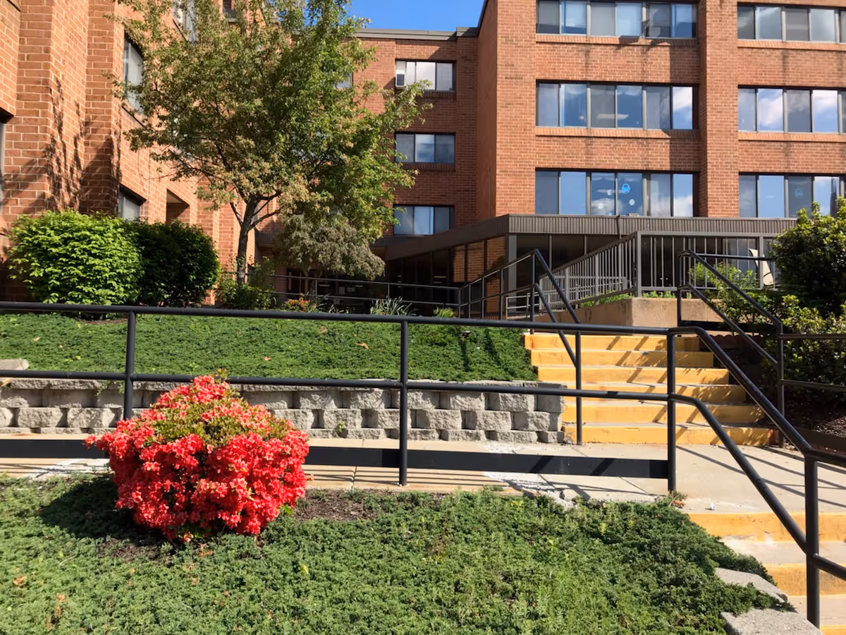 Brick multi-story building entrance with yellow steps, metal handrails, a concrete ramp and landscaped greenery including a red flowering bush.