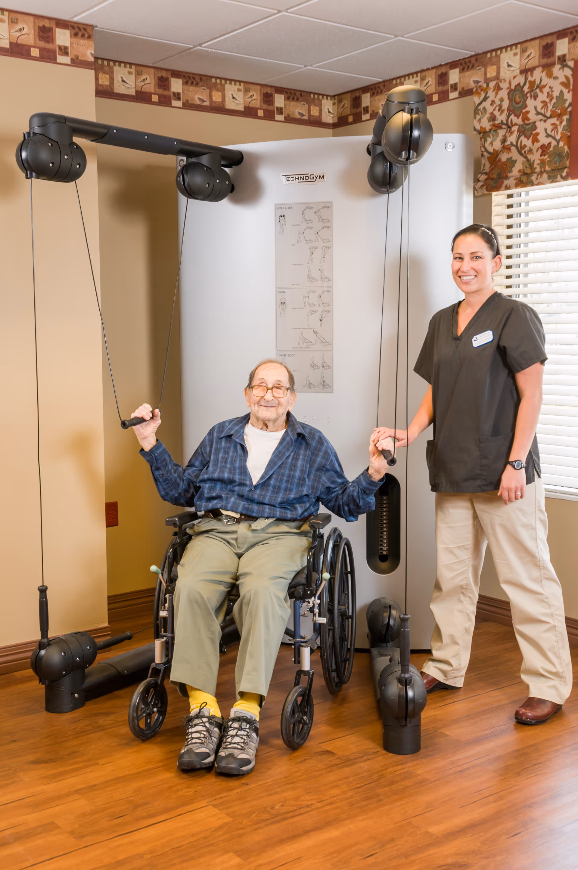 An elderly man in a wheelchair using a cable exercise machine with the assistance of a female caregiver standing beside him in a room with wooden flooring and a window with blinds.