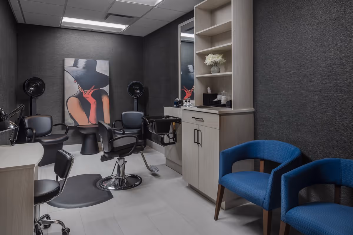A modern hair salon room with black salon chairs, a hair washing station, a large mirror, and a cabinet with shelves holding a vase with white flowers and hair care products. The walls are dark textured, and there is a stylish painting of a woman wearing a large black hat and red gloves. Two blue upholstered chairs are placed against the right wall.