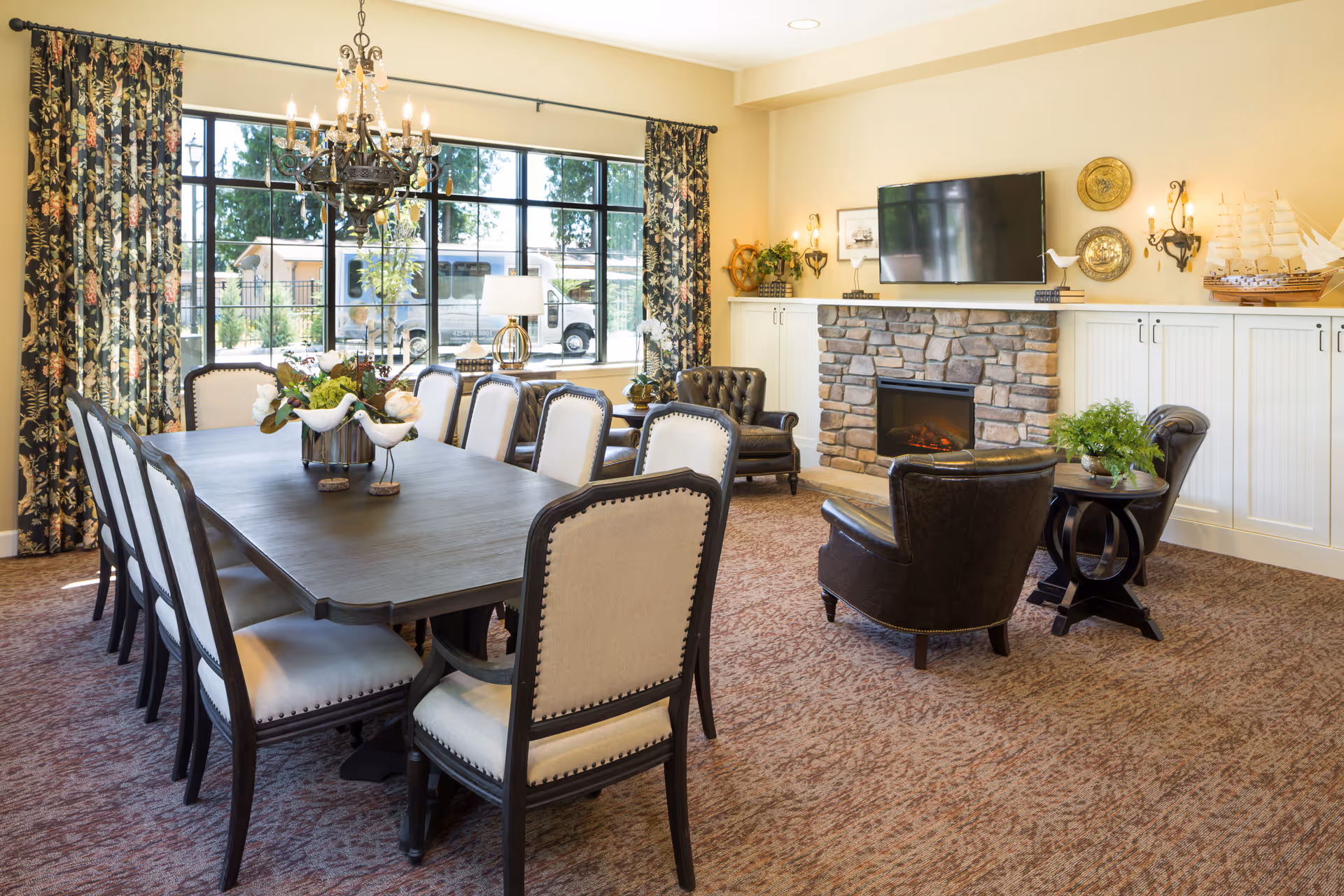 A well-lit, elegant common area featuring a long dining table with eight upholstered chairs, floral curtains on large windows, a stone fireplace with a mounted flat-screen TV above it, two leather armchairs with a small round table between them, and decorative items including a model ship and wall sconces.