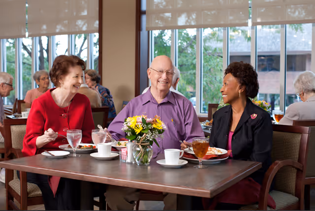 Three elderly people sitting at a dining table in a well-lit room with large windows. They are smiling and enjoying a meal together. The table has plates of food, glasses of water and iced tea, coffee cups, and a vase with yellow flowers. Other people are visible in the background also seated and dining.