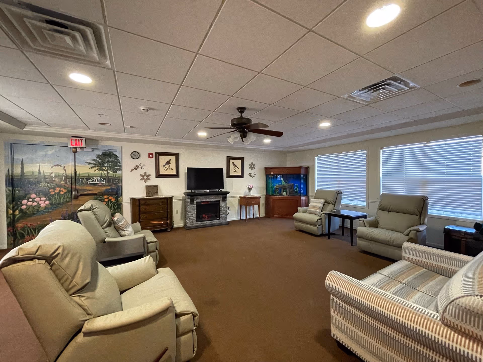 A cozy living room with several cushioned armchairs arranged around a central area. The room features a ceiling fan with lights, a flat-screen TV mounted above an electric fireplace, a wooden cabinet with an aquarium, and a mural painted on the door depicting a garden scene. Large windows with blinds allow natural light to enter the space.