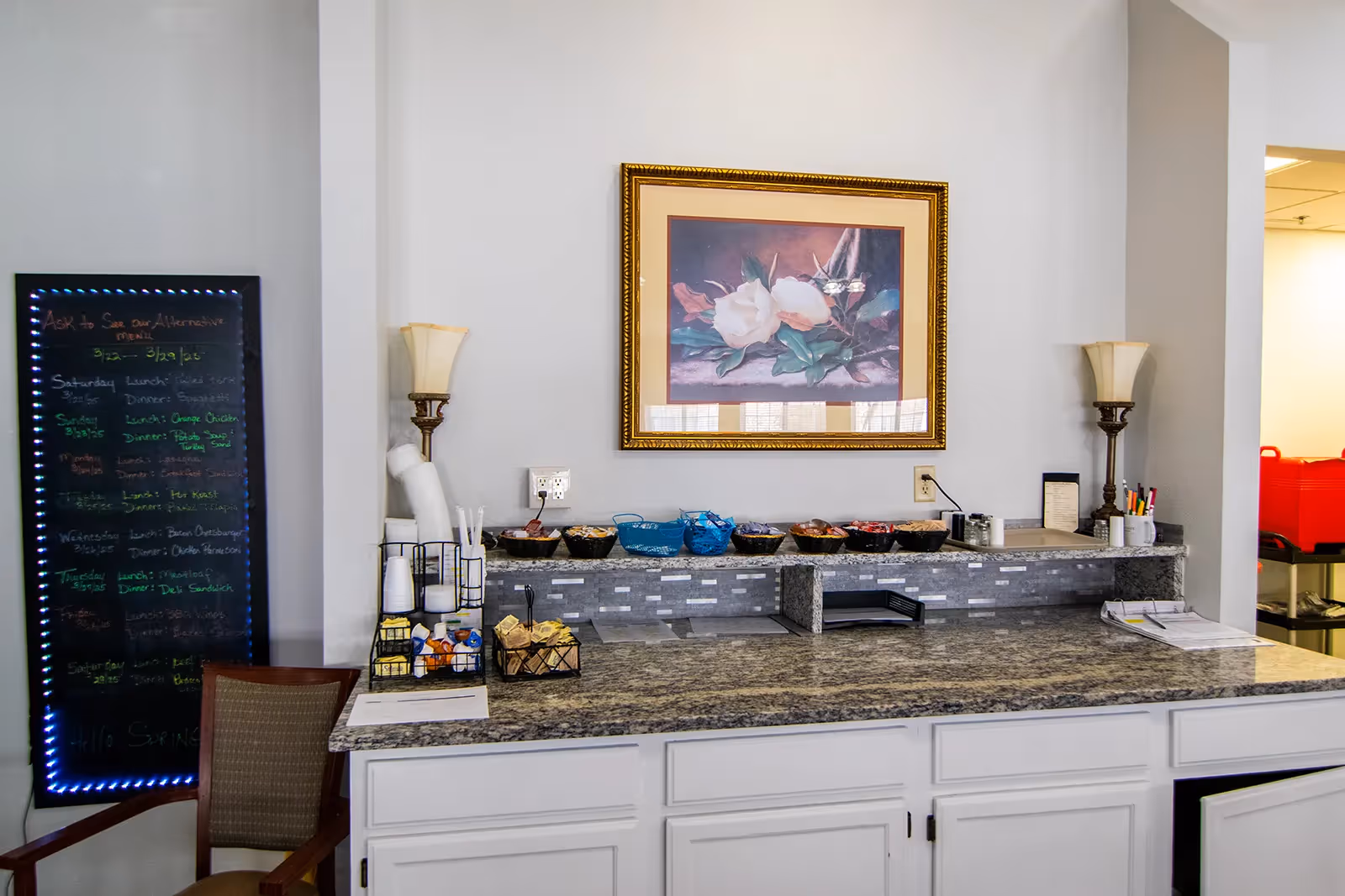 A countertop area with a granite surface and white cabinets below. On the counter are various bowls with snacks, cups, and condiments. Two decorative lamps flank a framed floral painting on the wall above the counter. To the left, there is a chair and a blackboard menu with colorful writing. The area appears to be part of a dining or snack service space.
