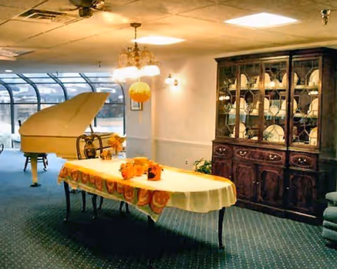 A room with a long table covered with a white tablecloth and decorated with orange-themed items. To the left, there is a white grand piano near large windows with a curved design. On the right side, there is a dark wooden china cabinet filled with plates and other dishware. The room has carpeted flooring and ceiling lights.