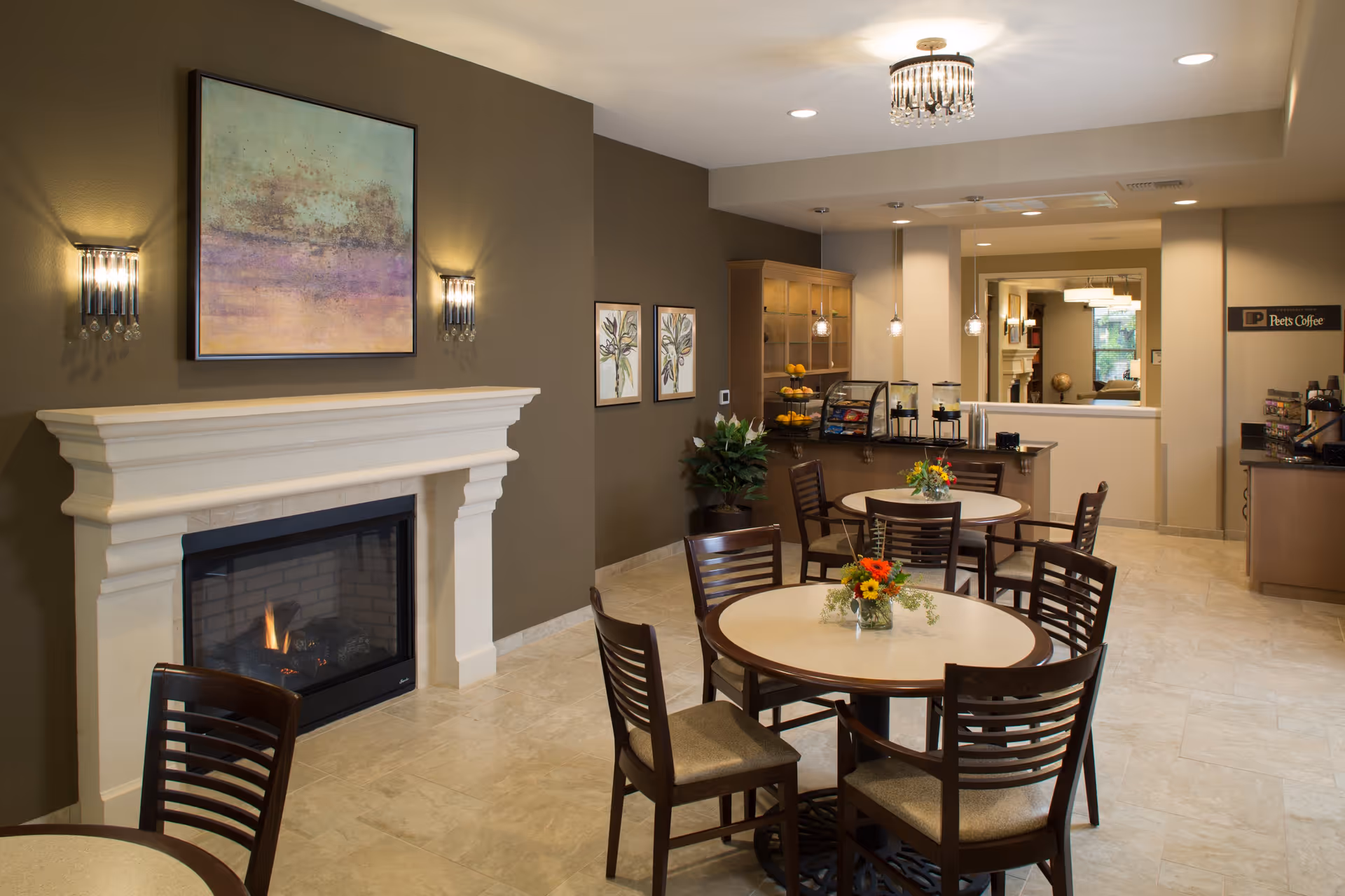 A cozy common area with round tables and chairs, a lit fireplace with a white mantel, wall sconces, and artwork above the fireplace. In the background, there is a counter with a display case containing snacks and beverages, a coffee station labeled 'Peet's Coffee,' and pendant lights hanging from the ceiling.