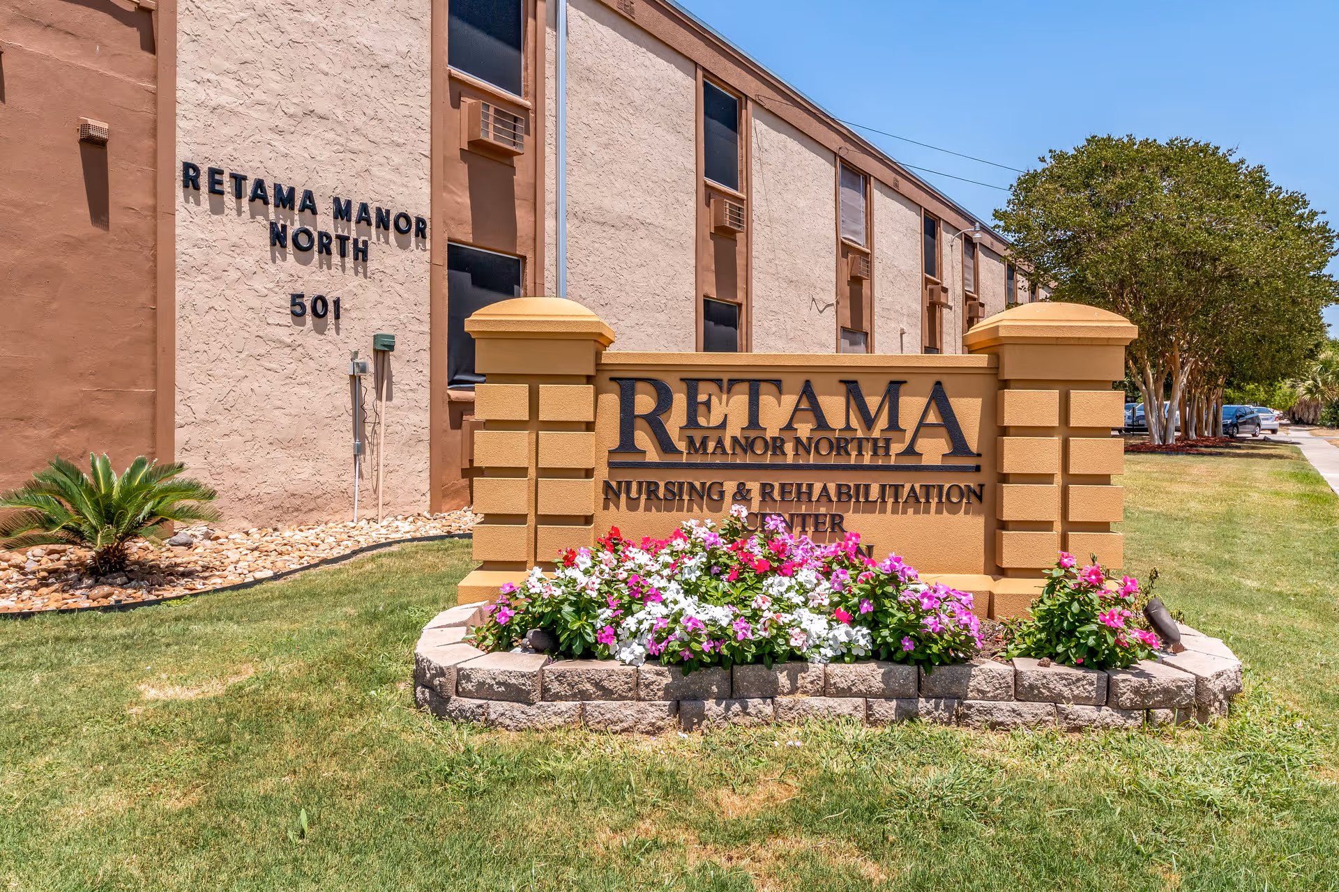 Stone sign reading 'RETAMA MANOR NORTH Nursing & Rehabilitation Center' with a flowerbed in front of a beige multi-story building.