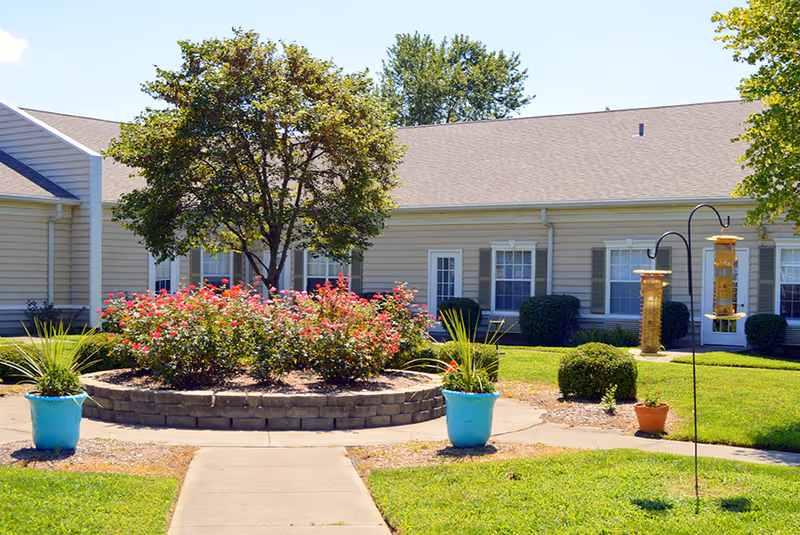 Outdoor courtyard area at Morning Pointe of Owensboro featuring a circular raised flower bed with blooming pink flowers and a tree in the center, surrounded by a paved walkway. There are two blue pots with green plants on either side of the walkway and bird feeders hanging on a metal stand on the right. The background shows a single-story building with beige siding, white doors, and windows with green shutters.