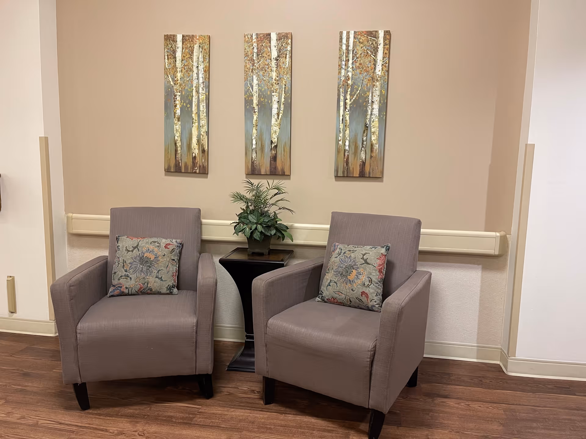 Two gray upholstered armchairs with patterned pillows flank a small table holding a potted plant beneath three vertical tree-themed paintings on a beige wall.