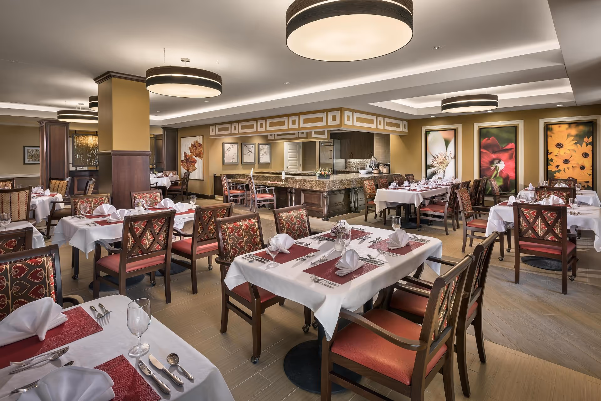 A spacious dining room with multiple tables set with white tablecloths, red placemats, folded napkins, silverware, and glasses. The room features wooden chairs with red and patterned upholstery, large circular ceiling lights, and decorative floral artwork on the walls. The overall ambiance is warm and inviting.