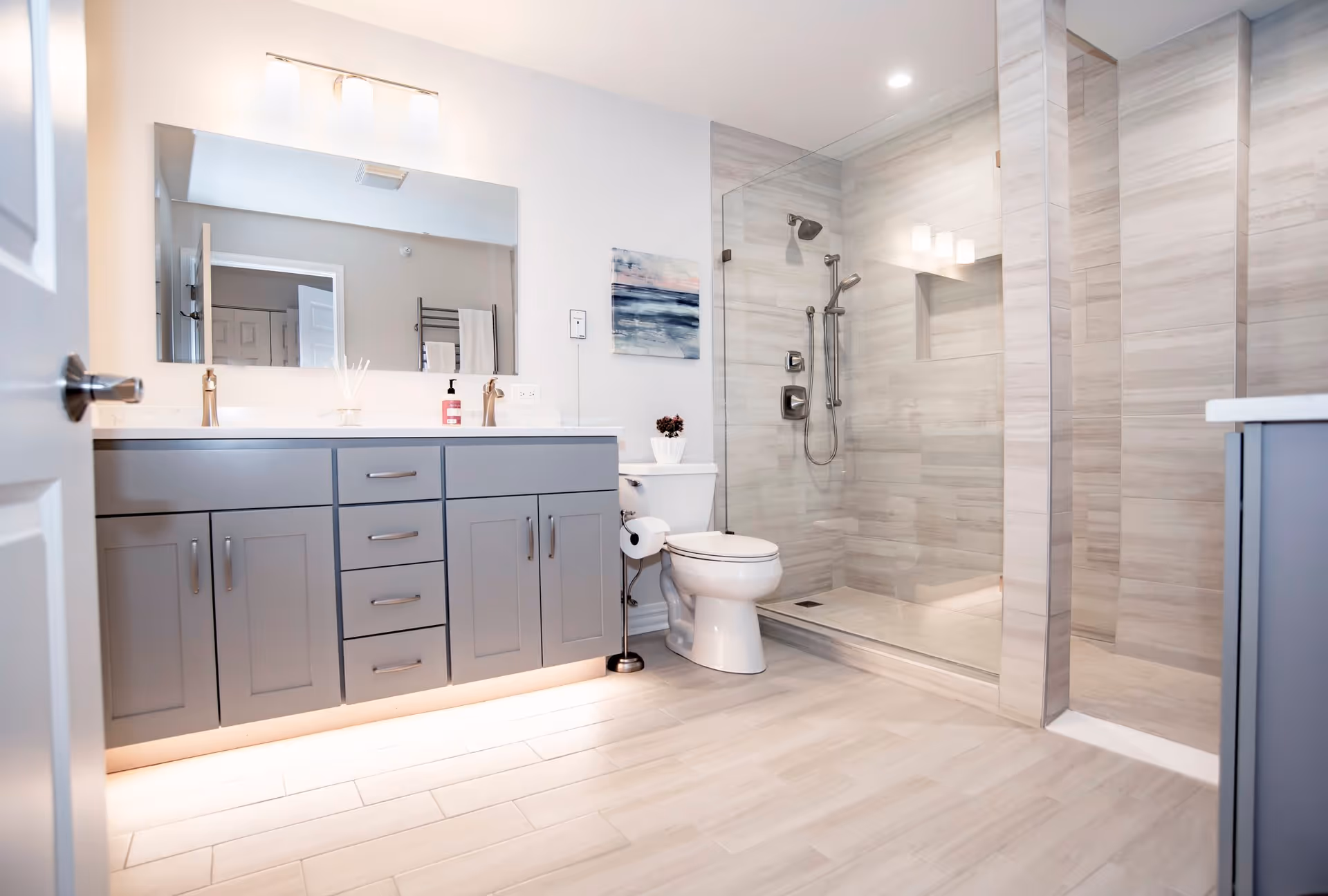 Modern bathroom with a large mirror above a gray vanity with multiple drawers and cabinets, a white toilet, and a spacious walk-in shower with glass doors and tiled walls. The bathroom has light-colored flooring and walls, with a towel rack and a piece of artwork on the wall.