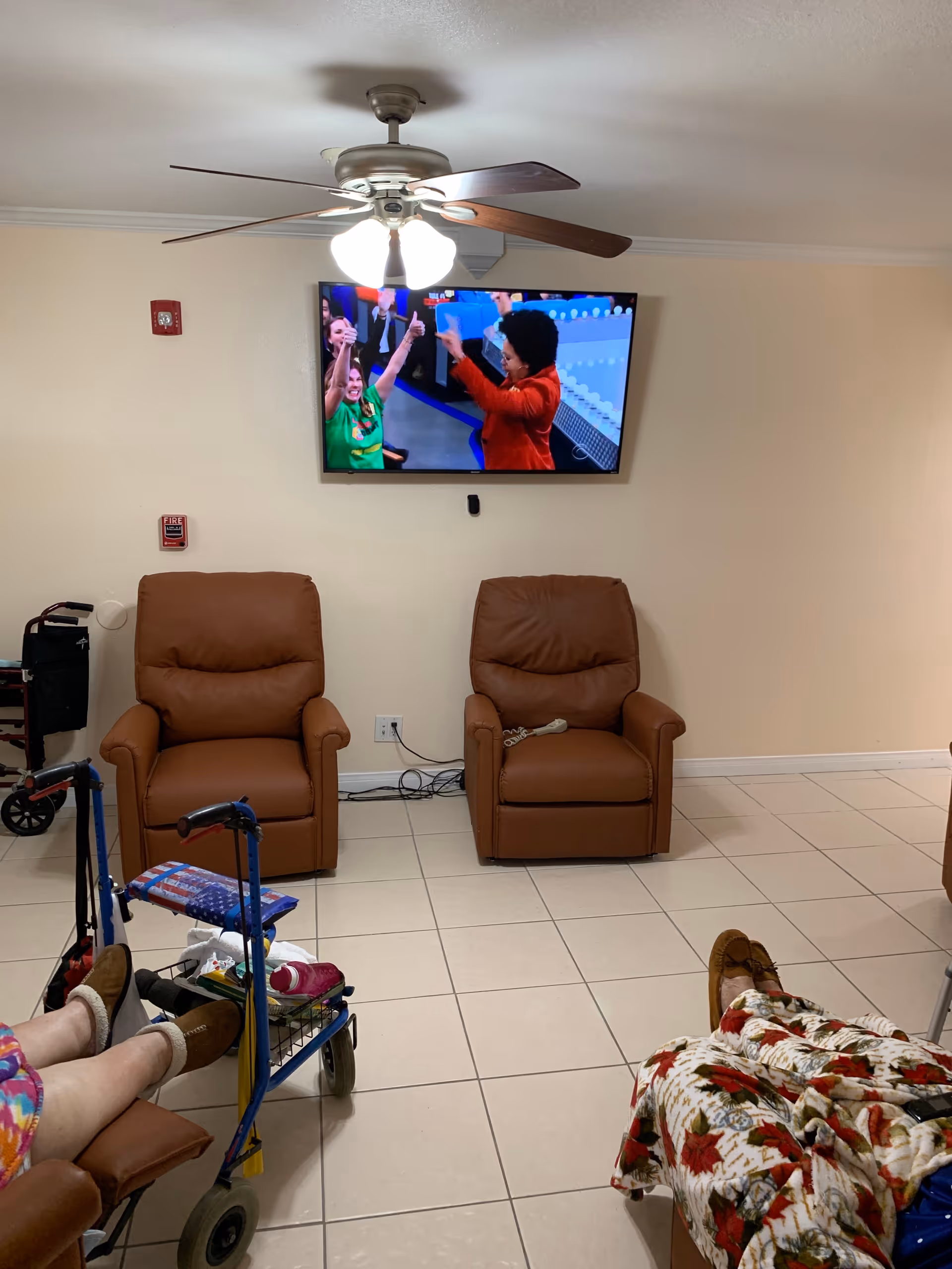 A room with two brown recliner chairs against a beige wall, a ceiling fan with lights above, and a flat-screen TV mounted on the wall showing people celebrating. Two elderly individuals are seated in the foreground with their legs visible, one using a walker with a basket containing personal items.