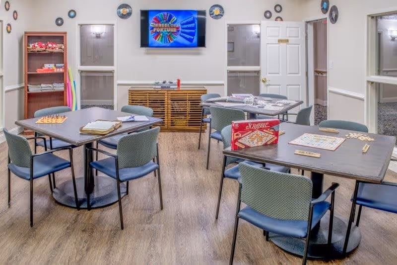 A senior living facility activity room with four tables and chairs arranged for games and socializing. The tables have board games such as Scrabble and chess set up. A TV mounted on the wall displays a colorful Wheel of Fortune game screen. The room has light-colored walls, wood flooring, and windows with white trim. A wooden cabinet is positioned below the TV, and a bookshelf with various items is visible on the left side of the room.