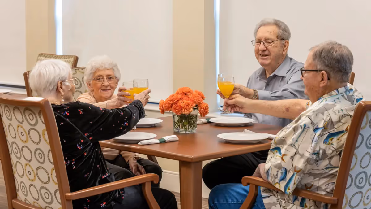 Four elderly residents seated around a dining table raising glasses of orange juice in a communal dining room.
