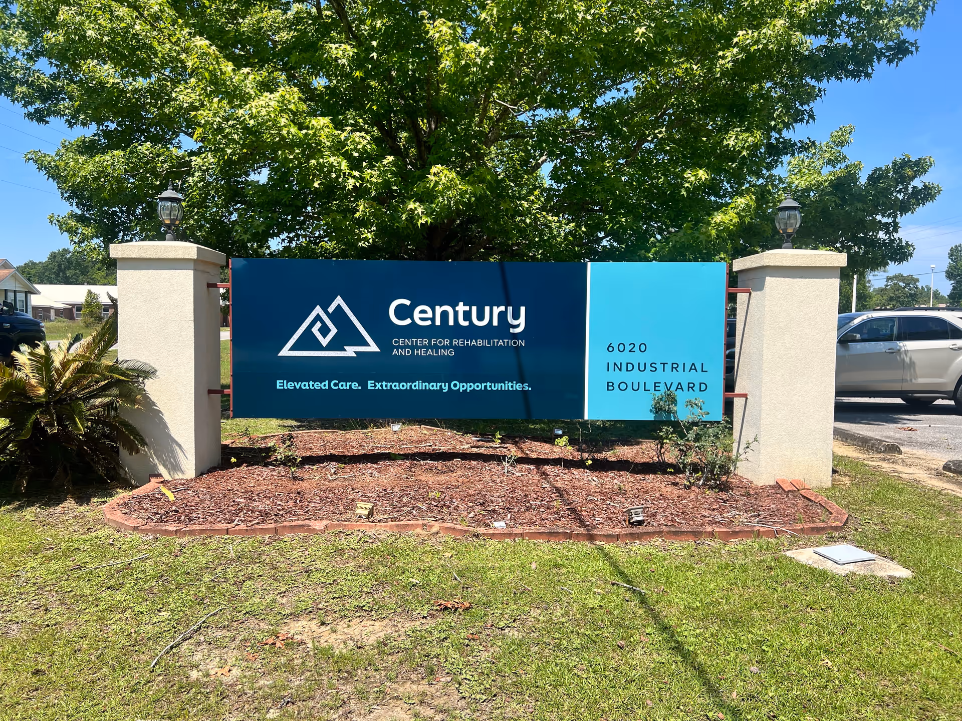 Outdoor view of a sign for Century Center for Rehabilitation and Healing, located at 6020 Industrial Boulevard. The sign is mounted between two white pillars with lamps on top, surrounded by mulch and greenery, with a tree and parked cars in the background under a clear blue sky.