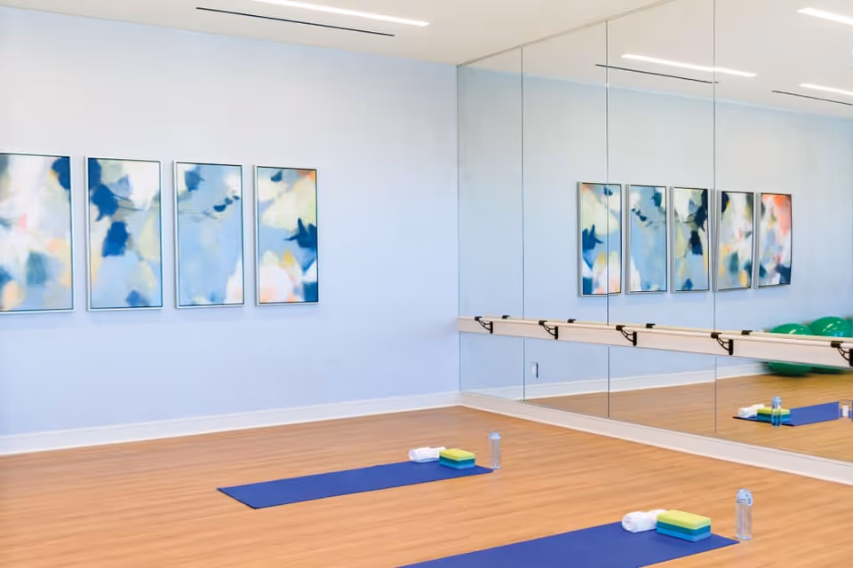 A bright exercise room with wooden flooring, two blue yoga mats laid out with towels, blocks, and water bottles on them. The room has a large wall mirror with a ballet barre and four abstract paintings on a light blue wall.