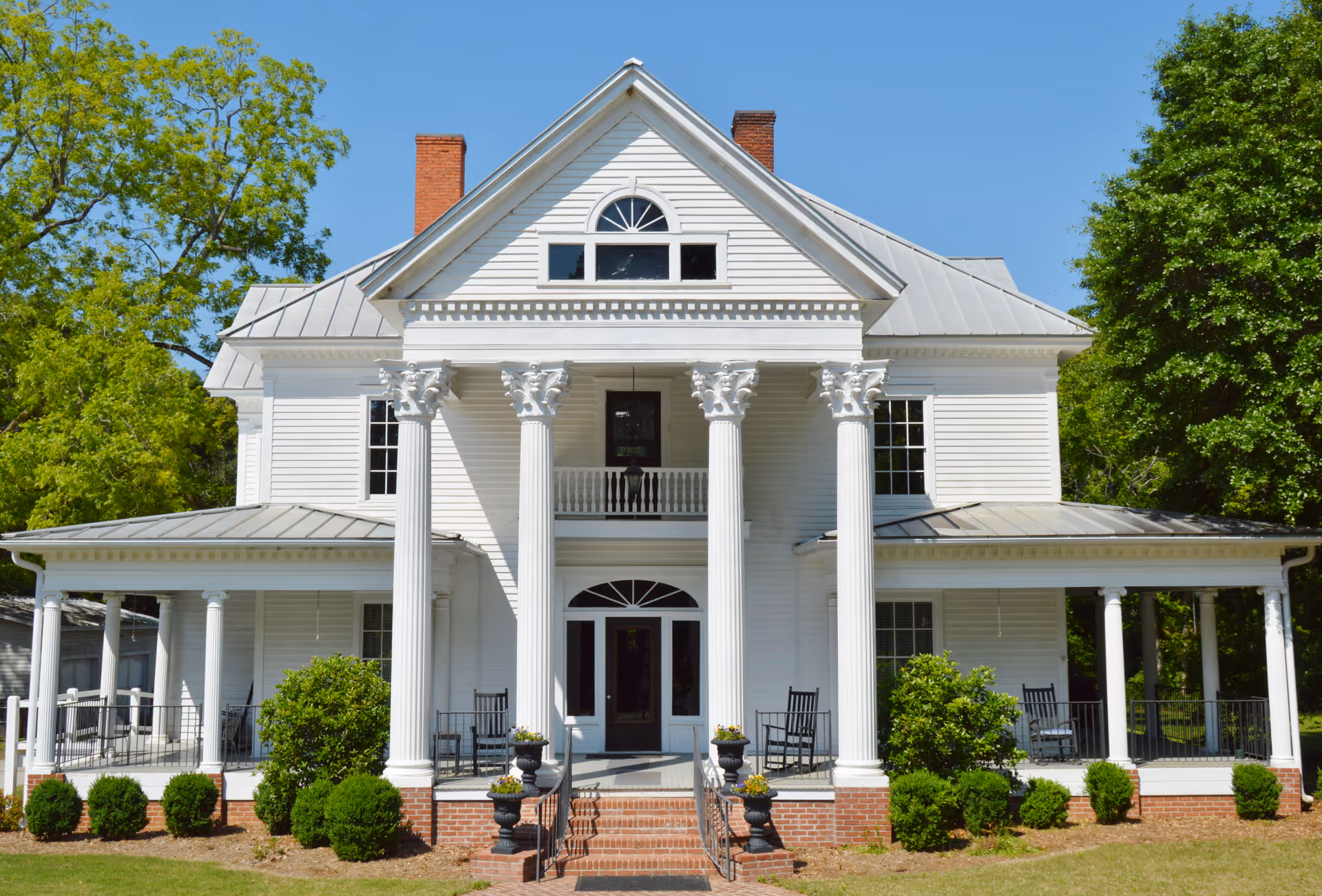Front exterior view of a large white two-story house with classical columns, a porch with rocking chairs, and greenery around the building under a clear blue sky.