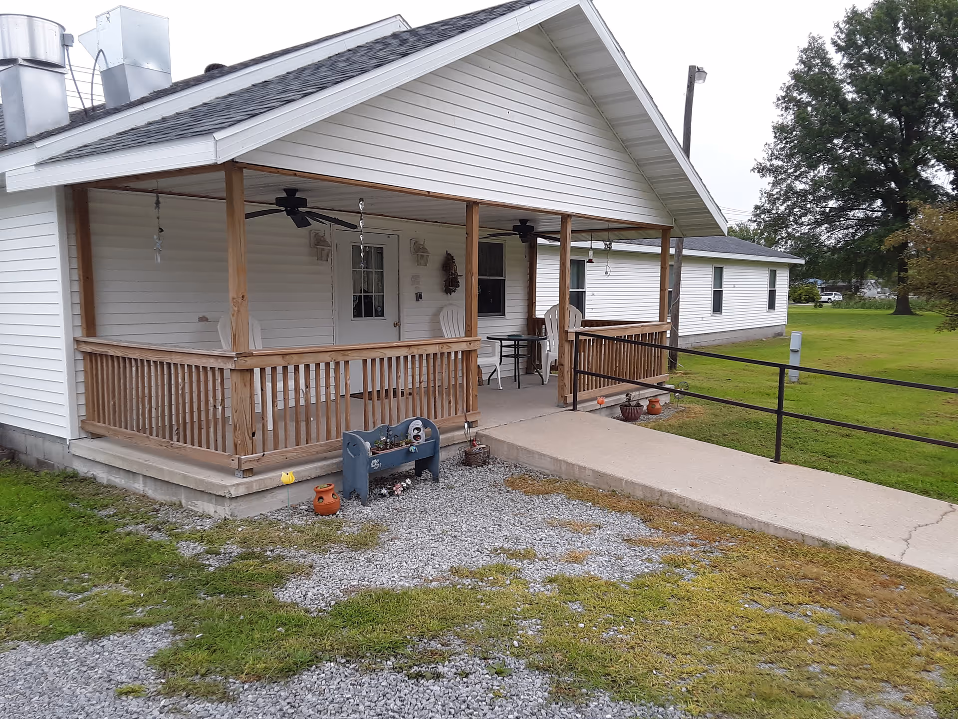 Covered wooden front porch and ramp of a single-story white house with chairs and a grassy yard.