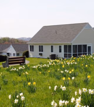 A grassy outdoor area with blooming yellow and white daffodils in front of a single-story light-colored building with a sloped roof. There is a wooden bench on the lawn and another building visible in the background under a clear sky.
