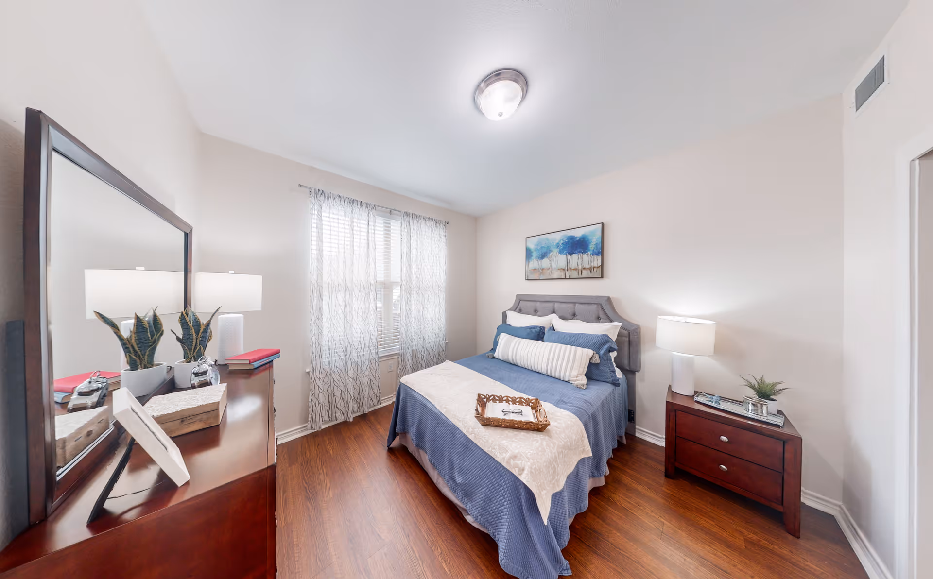 A neatly arranged bedroom with a bed covered in blue and beige bedding, a wooden nightstand with a lamp and small plant, a wooden dresser with a mirror, two potted plants, and a window with sheer curtains letting in natural light. A framed painting of trees hangs above the bed.