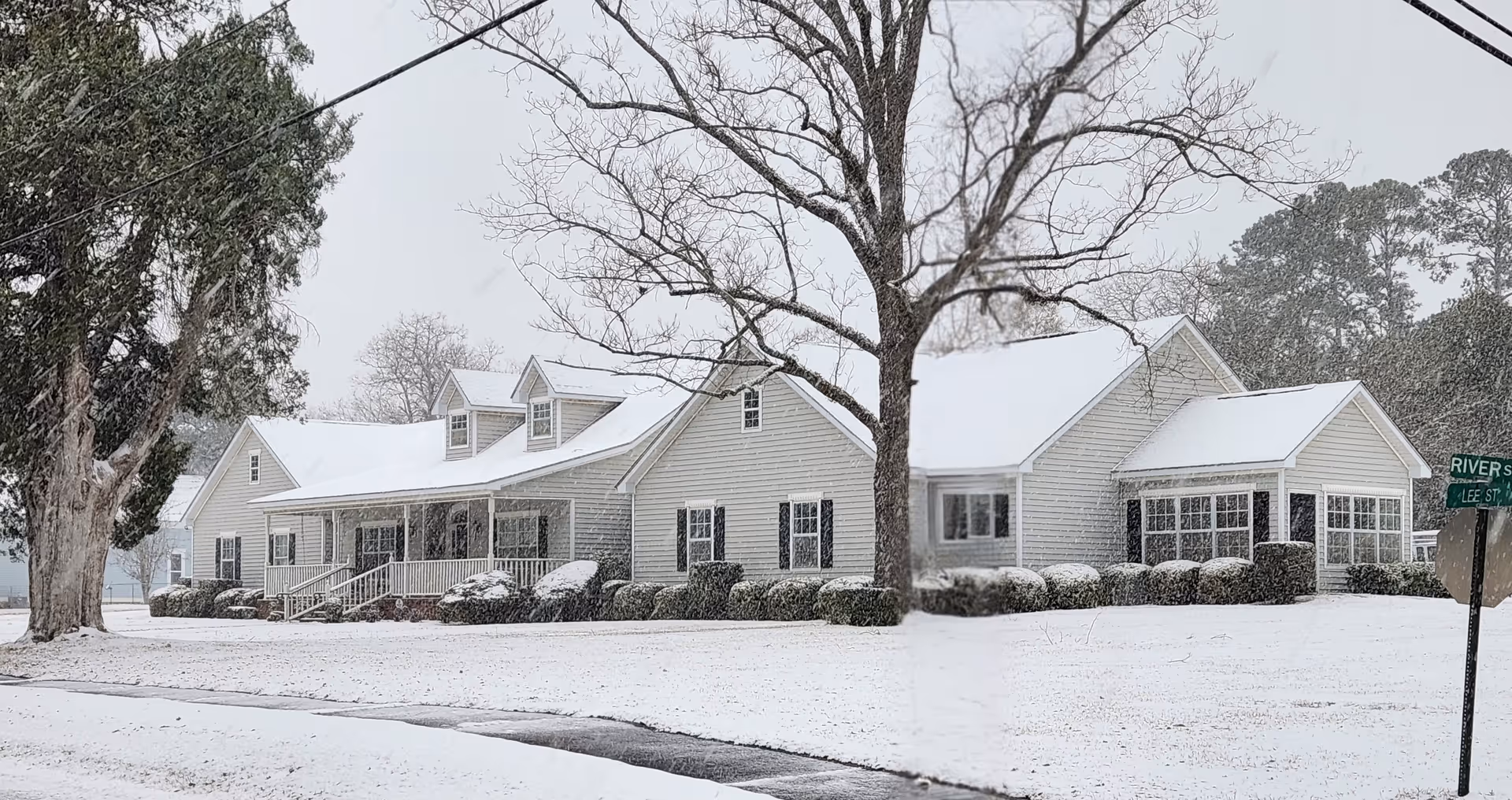 A large white building with a porch and multiple windows covered in a light layer of snow. There are leafless trees and bushes around the building, and snow covers the ground and roof. A street sign is visible on the right side showing River St and Lee St.