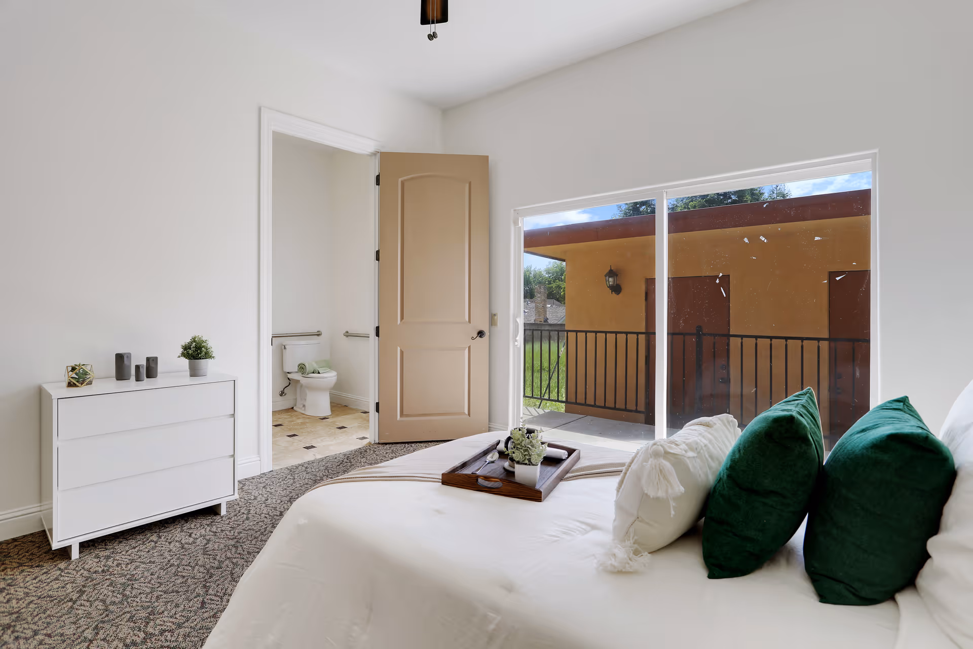 A bright bedroom with white bedding and green pillows, a dresser, an open doorway to a bathroom, and sliding glass doors opening to a balcony.
