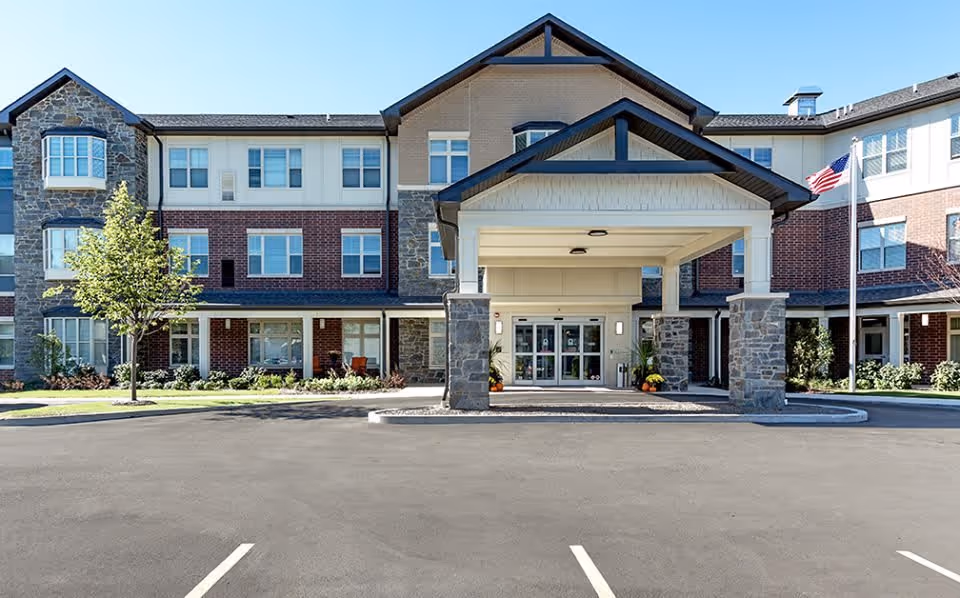 Front exterior view of a three-story senior living facility building with a covered entrance supported by stone pillars, a flagpole with an American flag, and a paved parking area in front.