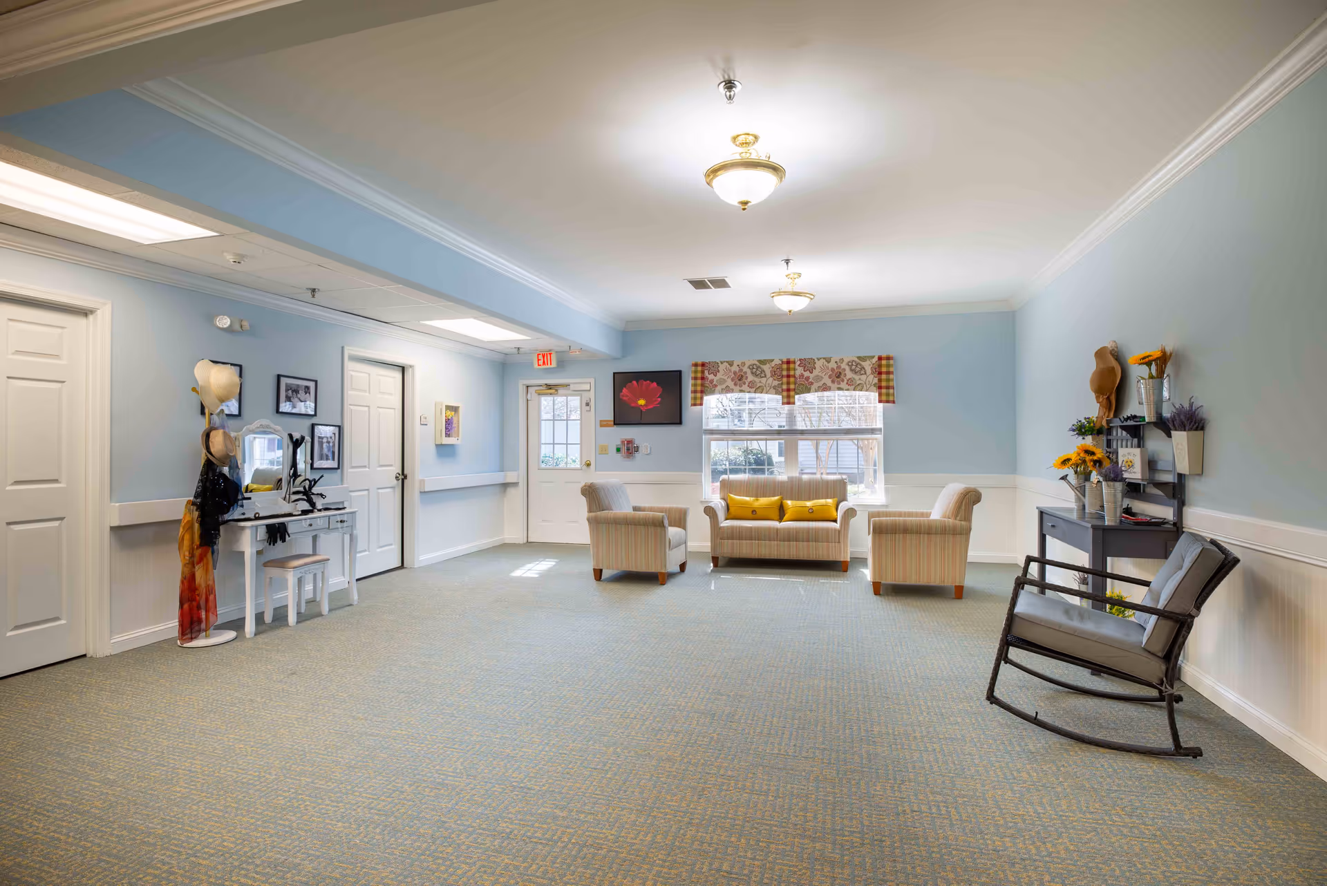 A spacious senior living common area with light blue walls and carpeted floor. The room features a small seating arrangement with a striped sofa and two matching armchairs, all with yellow accent pillows, positioned near a window with floral and plaid valances. There is a rocking chair with a cushion on the right side, a small table with decorative flowers and hats on the wall, and a white vanity table with a mirror and hats on a stand on the left side. The ceiling has two light fixtures, and there is an exit door at the back of the room.