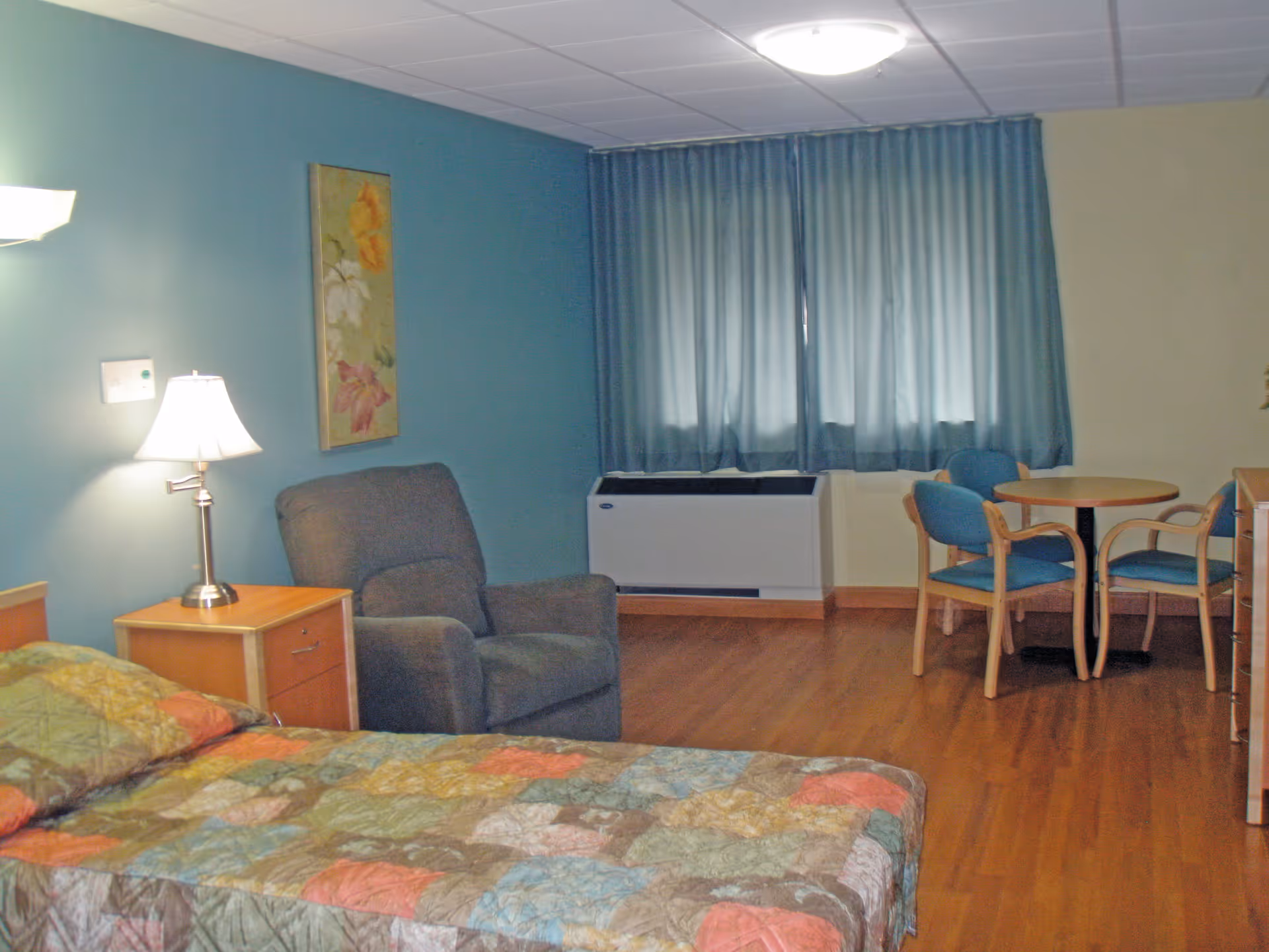 A senior living facility room with a bed covered in a colorful patchwork quilt, a wooden nightstand with a lamp, a gray upholstered armchair, a round wooden table with four blue cushioned chairs, blue curtains covering a window, and hardwood flooring.
